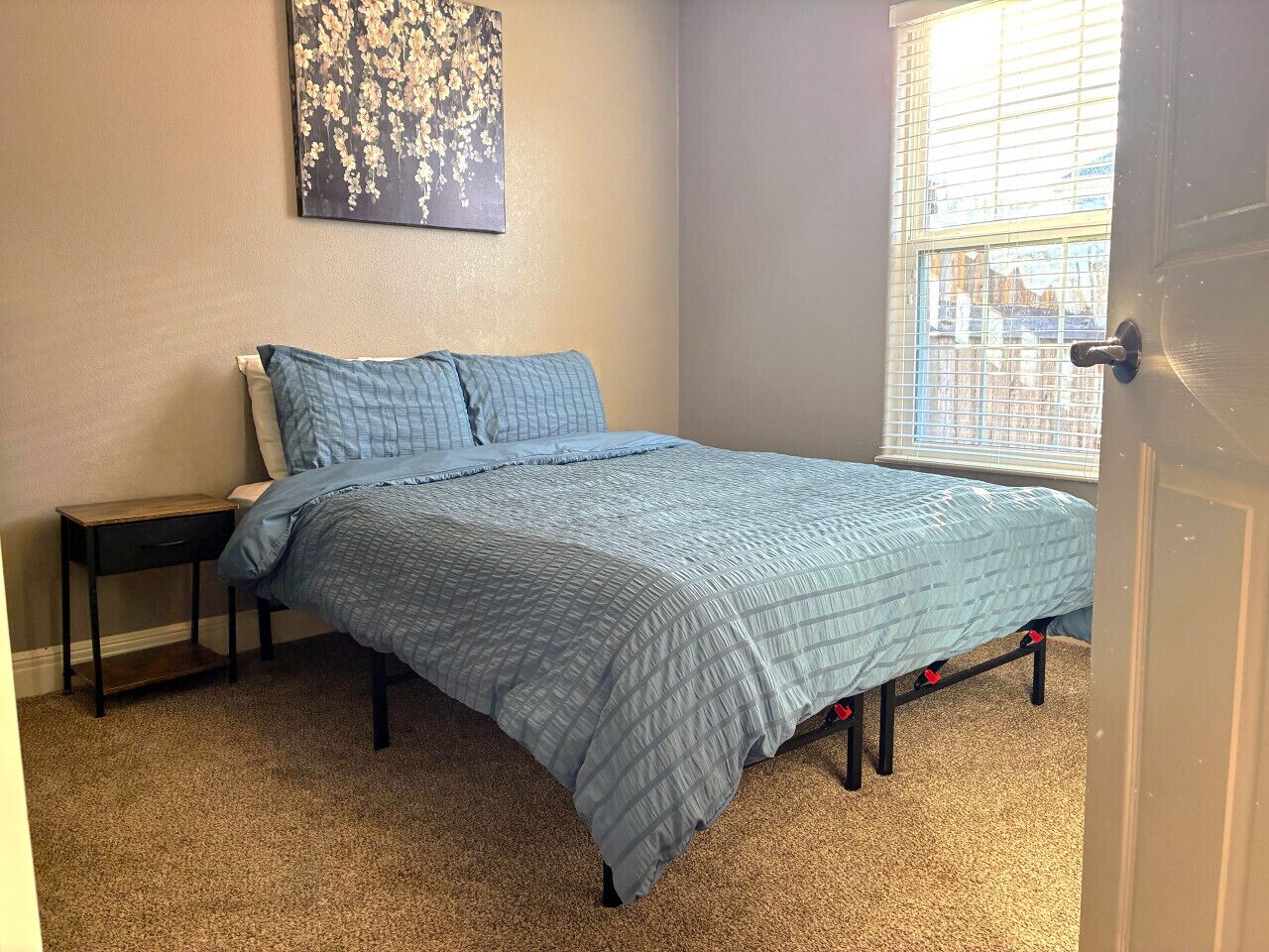 A bedroom with a bed, a nightstand, a window with blinds, and a wall-mounted artwork.