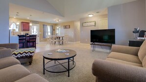 A spacious living room with a large TV, comfortable seating, a dining area, and a kitchen in the background.