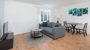 A modern living room with a grey sofa, a glass coffee table, a TV on a stand, a dining table with chairs, and a large window with curtains. - Spacious 1 Bedroom Apartment w/ Parking in Crawley (Crawley)