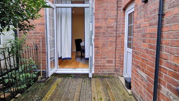 A wooden deck leading to a room with a white door, a brick wall, and a small table with chairs inside.