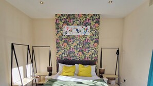 A neatly made bed with white linens and two yellow pillows, a floral tapestry on the wall, and a pair of black floor lamps on either side.