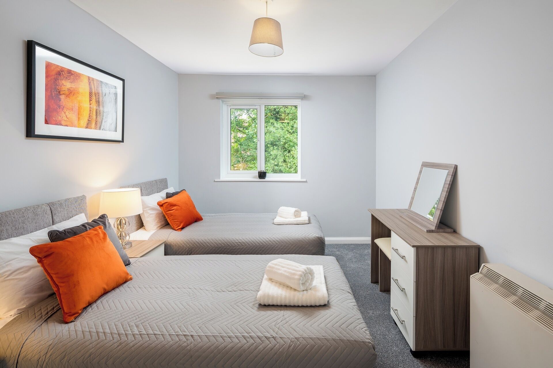 A modern hotel room with two beds, a desk, a window with a view of greenery, and a framed picture on the wall.