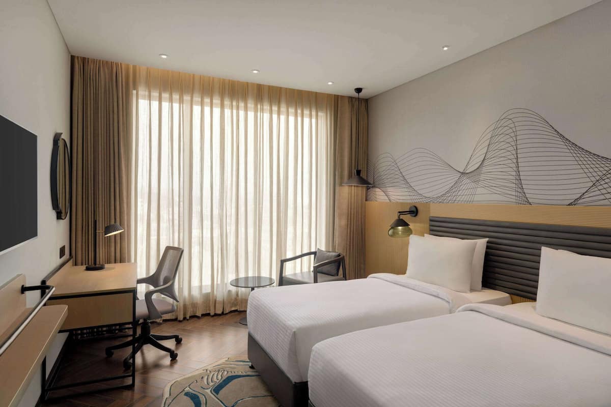 Ein Hotelzimmer mit zwei Betten, einem Schreibtisch, einem Stuhl, einem kleinen Tisch und einem großen Fenster mit Vorhängen.