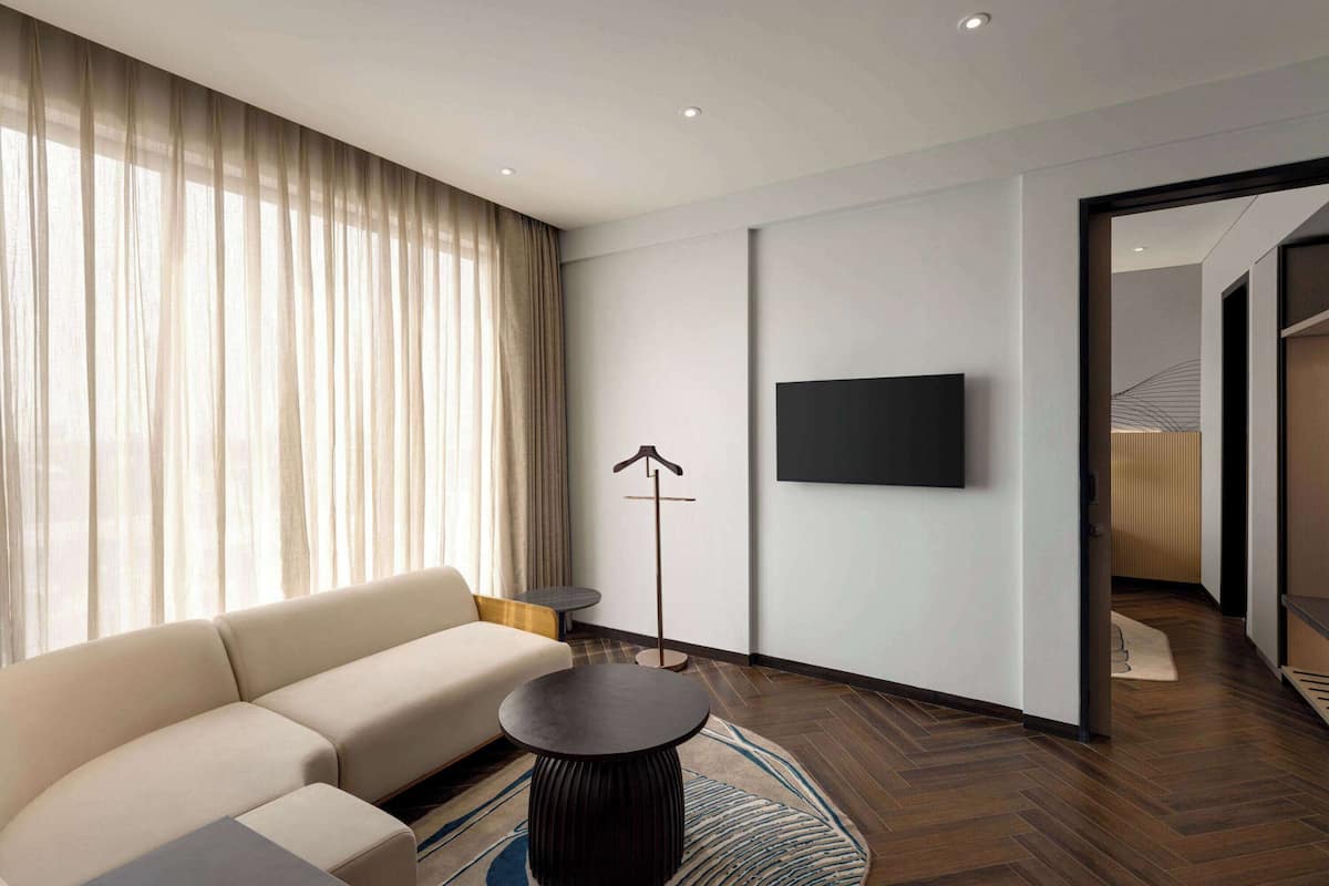 Ein modernes Hotelzimmer mit einer Couch, einem runden Couchtisch, einem flachbildfernseher an der Wand und einer Stehlampe.