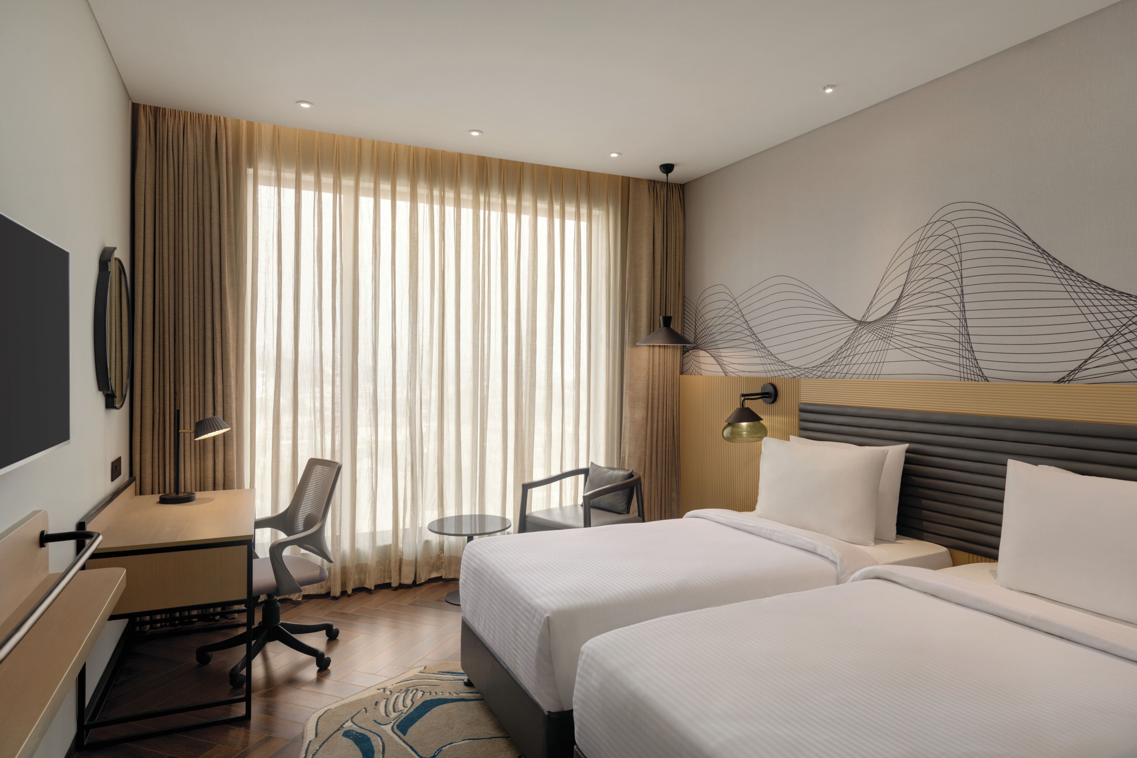 Ein Hotelzimmer mit zwei Betten, einem Schreibtisch, einem Stuhl, einem kleinen Tisch und einem an der Wand befestigten Fernseher.