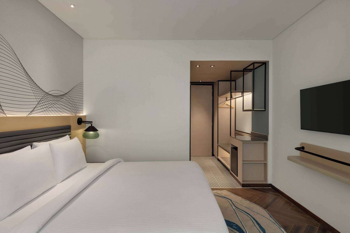 Ein modernes Hotelzimmer mit einem aufgeräumten Bett, einem an der Wand montierten Fernseher und einem begehbaren Kleiderschrank mit Spiegel.