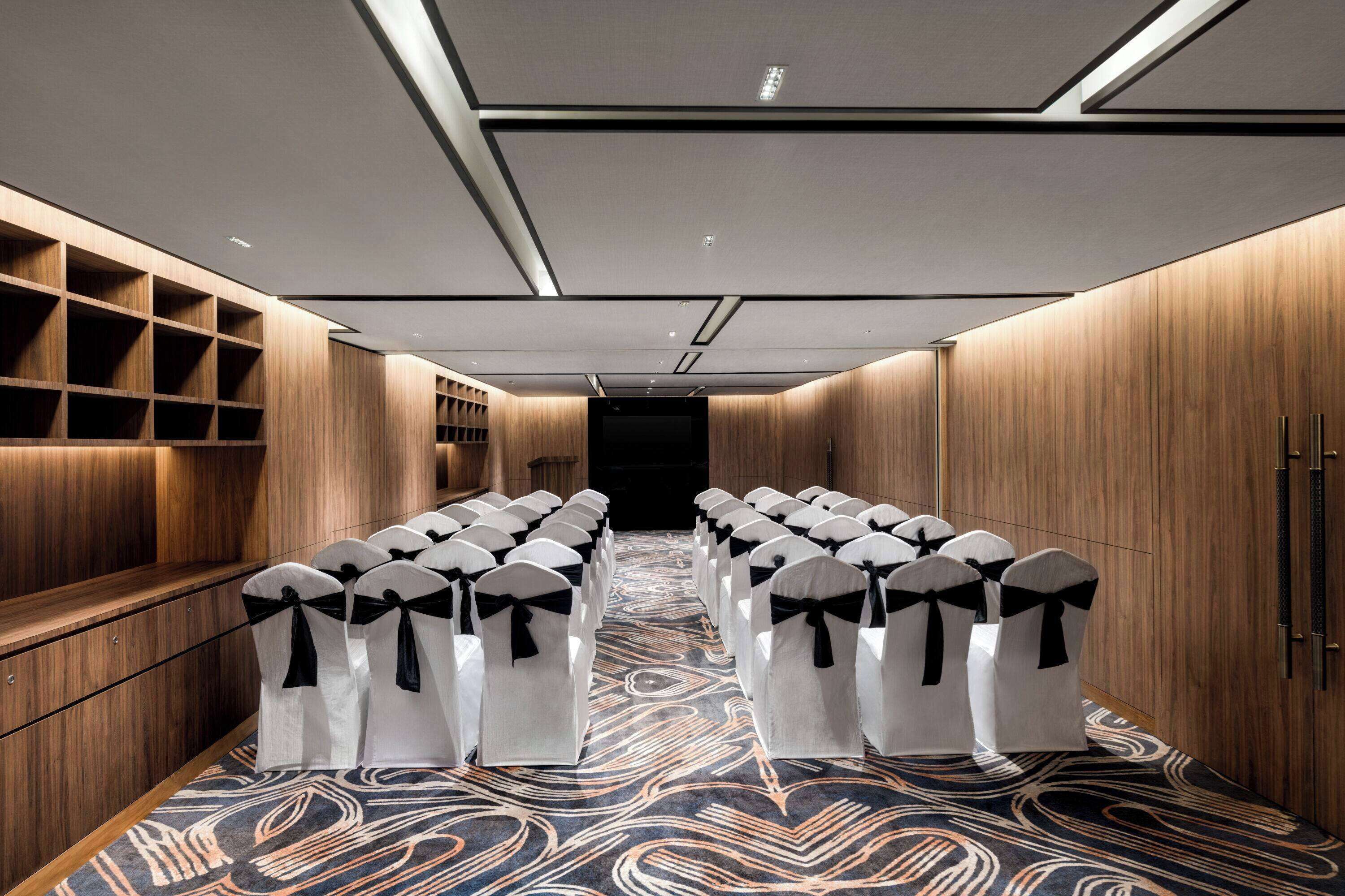 Une chambre d’hôtel avec une longue rangée de chaises blanches ornées de rubans noirs, des lambris en bois sur les murs et un tapis à motifs.