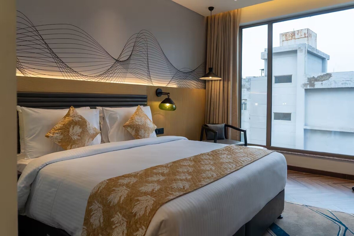 Een moderne hotelkamer met een groot bed, een hoofdeinde met een bijzondere lamp, uitzicht op de stad door grote ramen en een klein zithoekje.