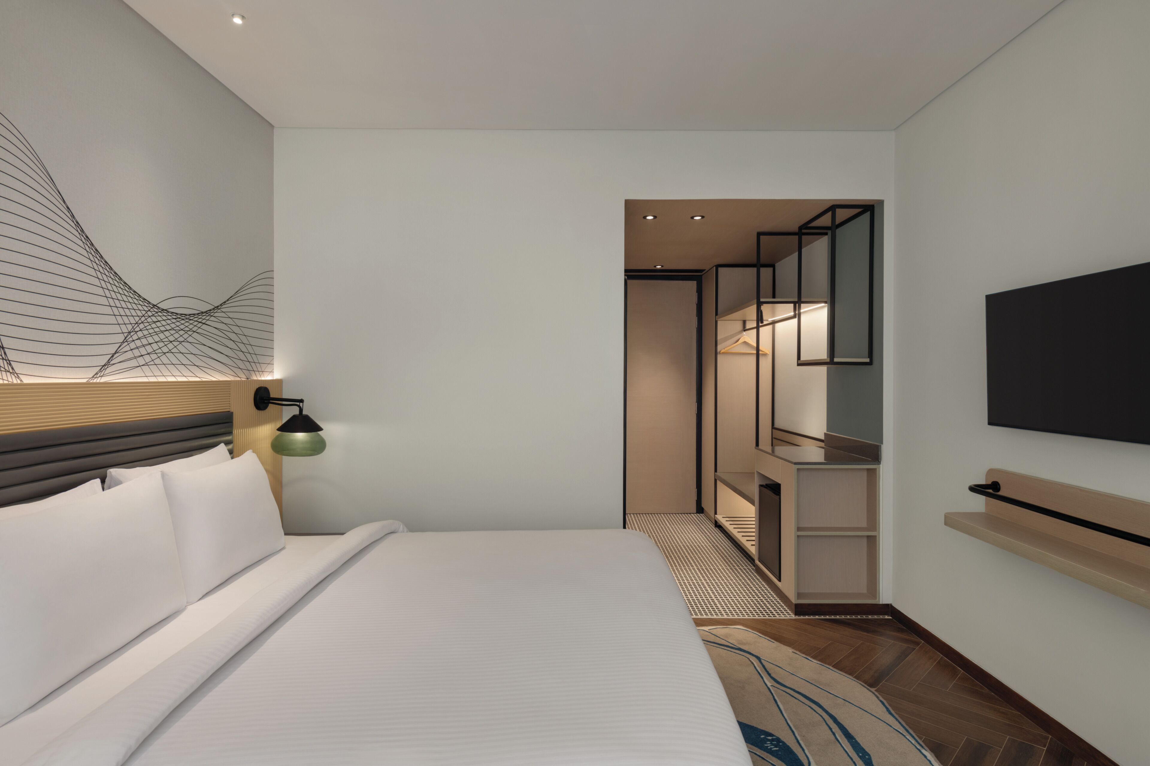 A modern hotel room with a bed, a wall-mounted TV, a bedside lamp, and a wardrobe.