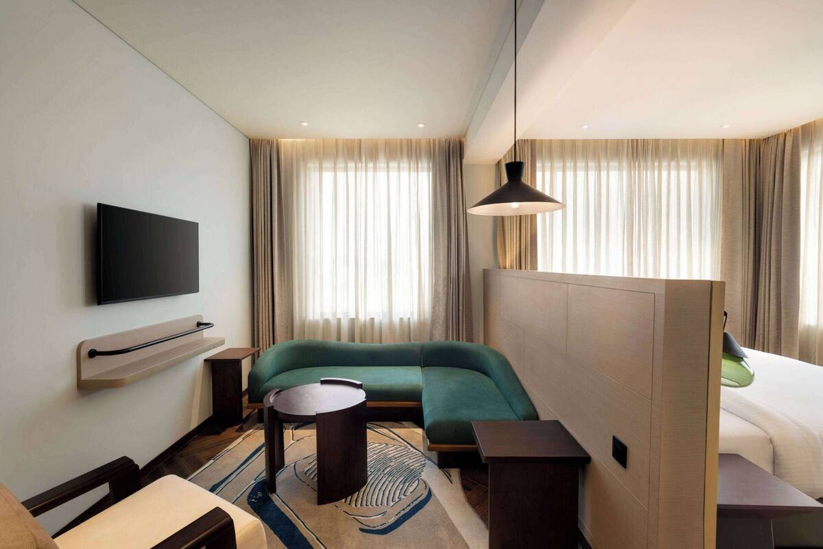 Ein Hotelzimmer mit einem wandmontierten Flachbildfernseher, einem grünen Sofa, einem kleinen runden Tisch, einem Bett mit einem grünen Kissen und einem großen Fenster mit Vorhängen.