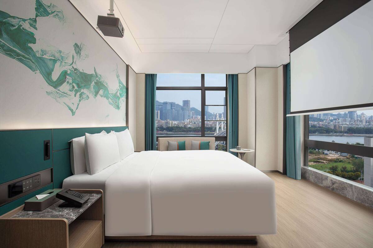 Une chambre d’hôtel moderne, dotée d’un grand lit, d’un bureau et offrant une vue sur le paysage urbain grâce à de grandes fenêtres.