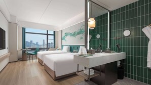 Ett modernt hotellrum med två sängar, ett stort fönster med utsikt över staden, ett skrivbord och ett badrum med gröna kakelplattor.