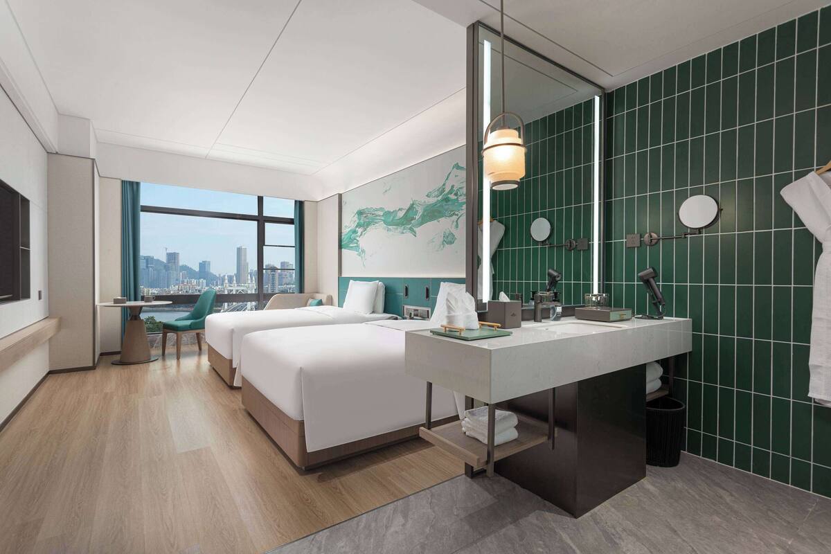 Une chambre d’hôtel moderne avec deux lits, une grande fenêtre donnant sur la ville, un bureau et une salle de bain aux carreaux verts.