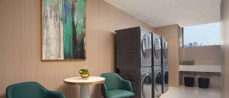 A modern room with a large painting, a washing machine, two chairs, a small table with a plant, and a view of the city.