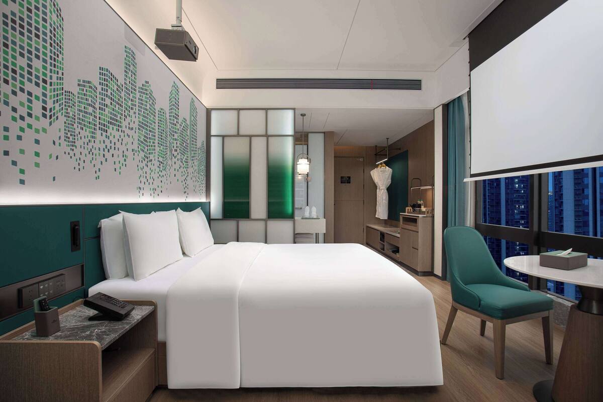 Une chambre d’hôtel moderne avec un grand lit, un bureau et une chaise. Un mur est décoré d’une fresque représentant un paysage urbain, il y a une fenêtre donnant sur les bâtiments et un grand miroir.