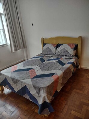 Un lit simple avec une tête de lit en bois, dans une pièce dont le sol est recouvert de planches de bois et qui possède une fenêtre avec des rideaux.