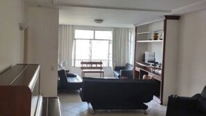 A living room with a sofa, a coffee table, and a TV stand. There is a window with curtains and a door leading to another room. - SUA CASA NO RIO-COPACABANA 3 QTS (Rio de Janeiro)