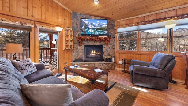 Ein gemütliches Blockhaus-Interieur mit Kamin aus Stein, Holzwänden, einem großen Fernseher und bequemen Sitzmöglichkeiten.