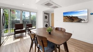 A modern dining area with a large table, chairs, and a potted plant. There is a large window with a view of greenery and a building. - Modern Gateway USC (Los Angeles)