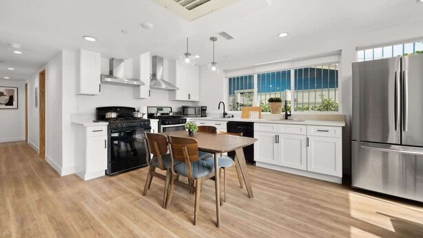 A modern kitchen with white cabinetry, a stainless steel refrigerator, and a dining area with wooden chairs. - Modern Gateway USC (Los Angeles)