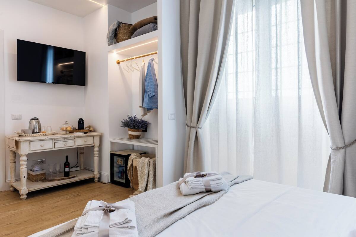 A hotel room with a bed, a TV mounted on the wall, a bedside table with a bottle, glasses, and a plant. There is a window with curtains and a shelf with towels and a basket.