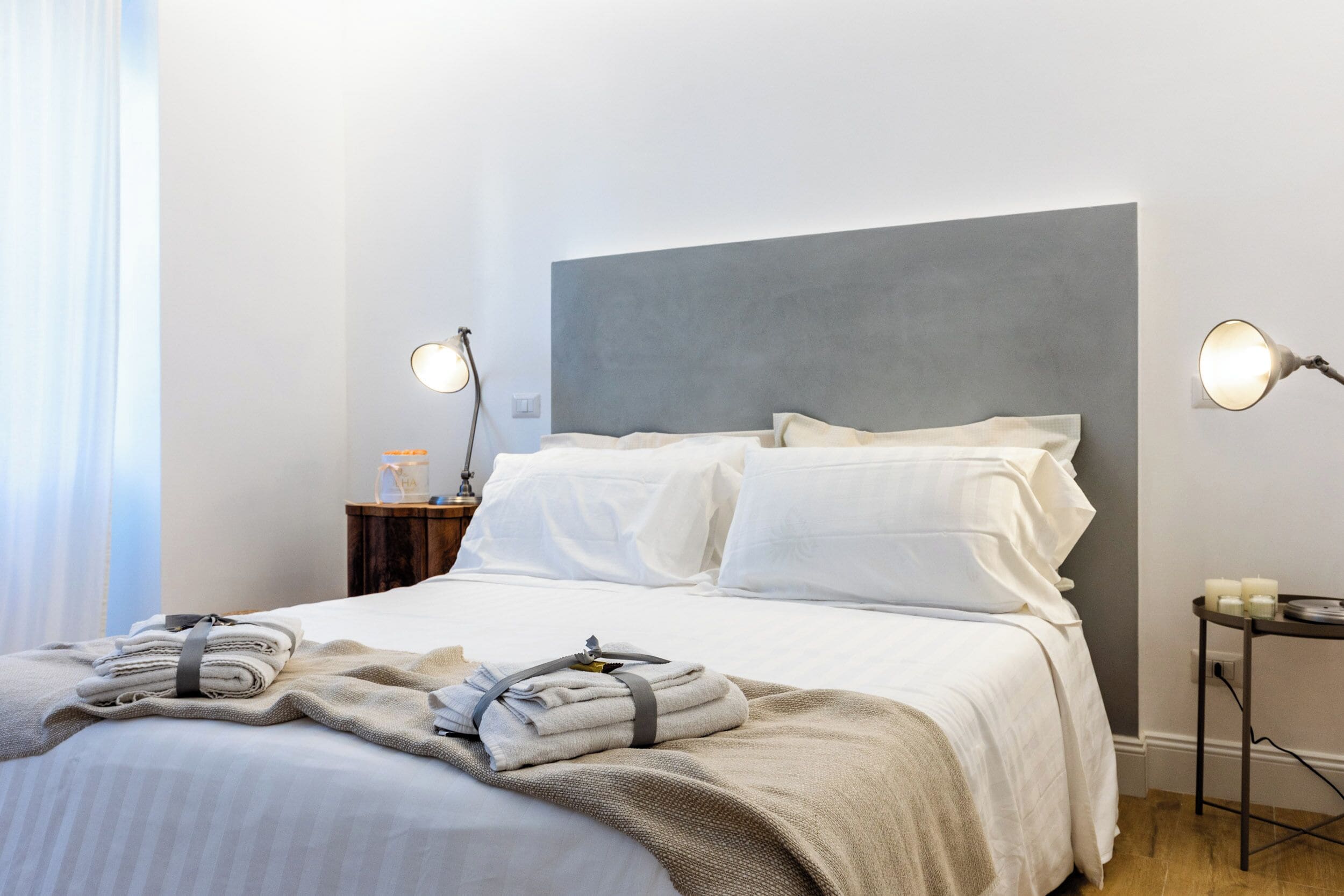 Una cama bien hecha con sábanas blancas y una manta color beige, una mesita de noche con una lámpara y otra mesita con lámpara y un pequeño adorno.