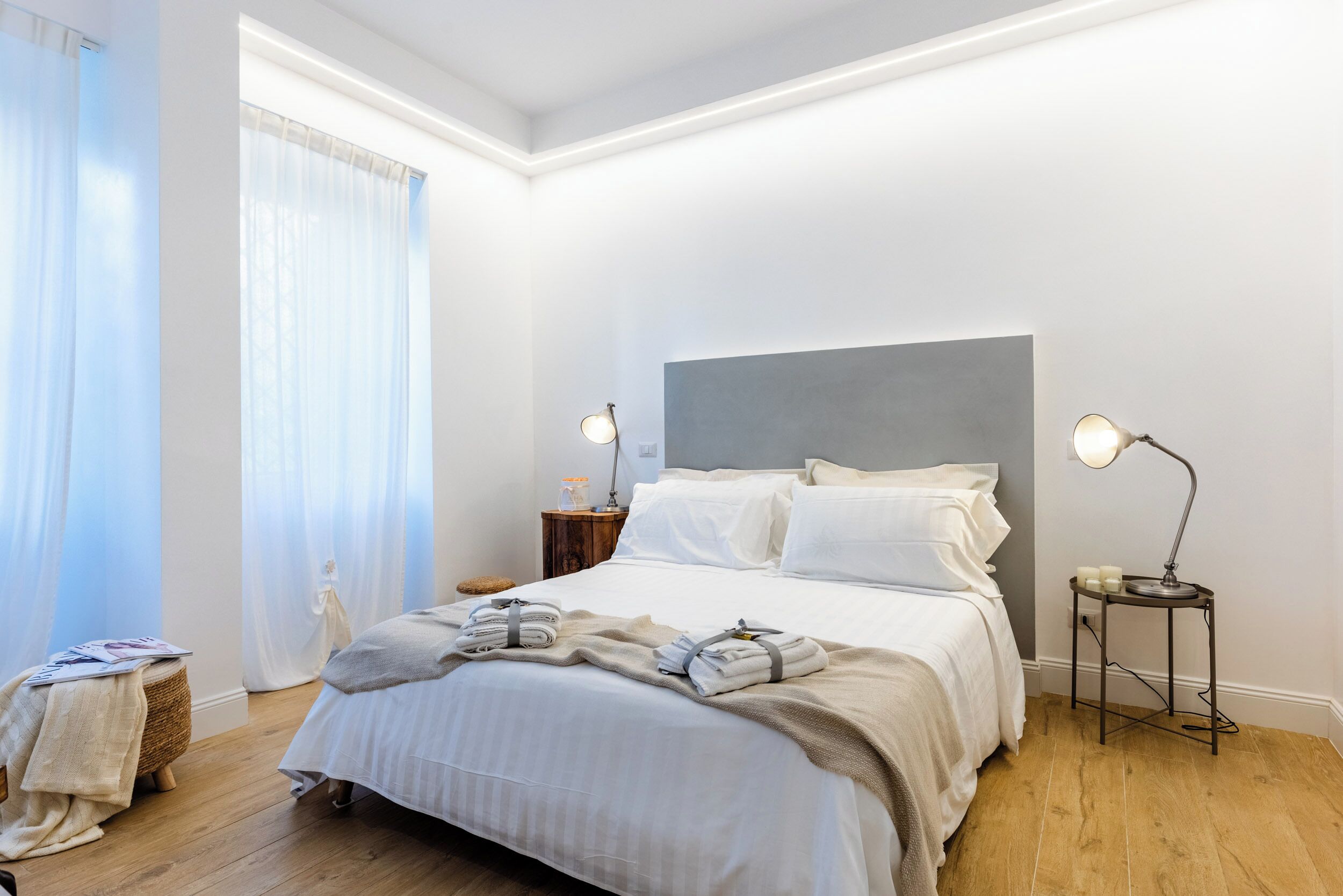 A neatly made bed with white linens, a grey headboard, and a bedside table with a lamp.