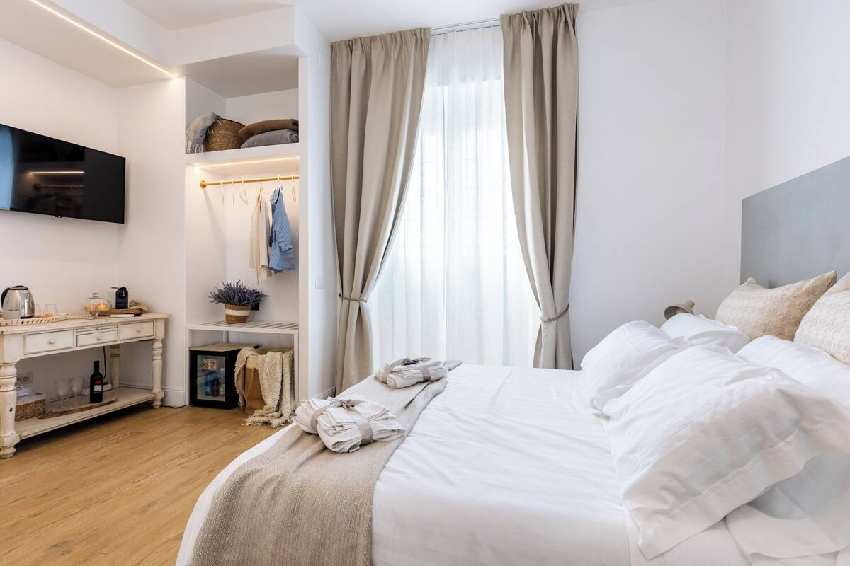 A modern bedroom with a large bed, a bedside table, a TV mounted on the wall, and a window with curtains.