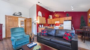 A living room with a blue armchair, a black leather sofa, and a wooden coffee table. A kitchen with wooden cabinets and a white refrigerator is visible in the background.