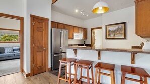 A modern kitchen with wooden cabinets, a stainless steel refrigerator, a microwave, and a large window. There are three wooden stools at the counter. - Stylish Mountain Retreat • Near RMNP & Downtown (Estes Park)