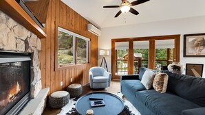 A cozy living room with a stone fireplace, a ceiling fan, a blue sofa, and a view of the outdoors. - Stylish Mountain Retreat • Near RMNP & Downtown (Estes Park)
