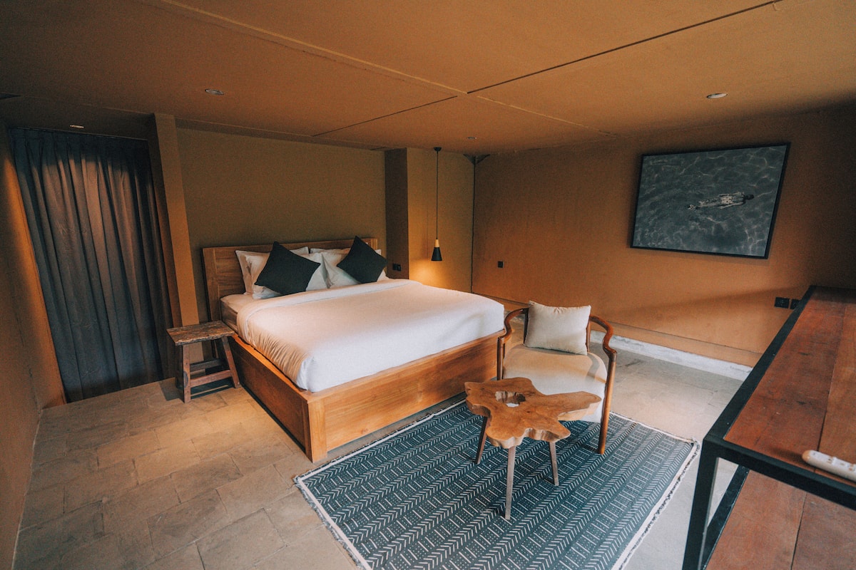 A bedroom with a wooden bed, a small wooden table, a chair, a patterned rug, and a painting on the wall.