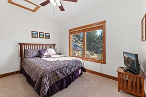 Quarto com cama, televisão em suporte de madeira, janela com vista para pedras e árvores, e ventilador de teto.