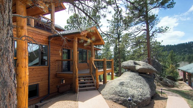 A wooden cabin with a balcony, large windows, and a stone feature in the yard.