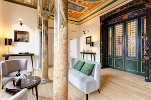 Une pièce luxueuse avec des colonnes en marbre, un canapé vert agrémenté de coussins verts, une table ronde avec une bouteille et des verres, et une porte décorative ornée de motifs circulaires.