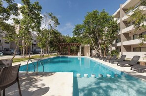 Une piscine dotée de chaises longues et d’une pergola, entourée d’arbres et d’un immeuble à plusieurs étages.