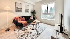 A modern living room with a black sofa, a round coffee table, a floor lamp, and a large window with a view of trees. - Stylish Downtown LA Gateway (Los Angeles)