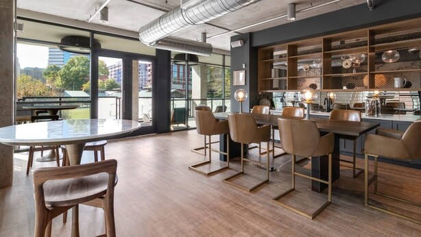A modern kitchen with a central island, wooden chairs, and a large window overlooking a cityscape. - Landing Apartments Durham (Durham)