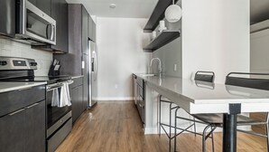 A modern kitchen with stainless steel appliances, a white countertop, and a dining area with a metal chair. - Landing Apartments Durham (Durham)