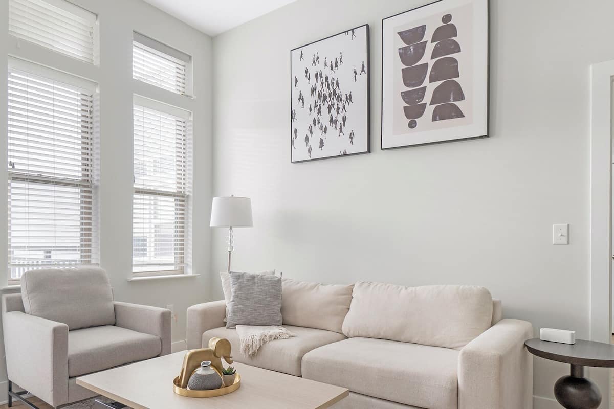 A modern living room with a beige sofa, a matching armchair, and a coffee table with a decorative item. There are two framed artworks on the wall, and a lamp is visible.