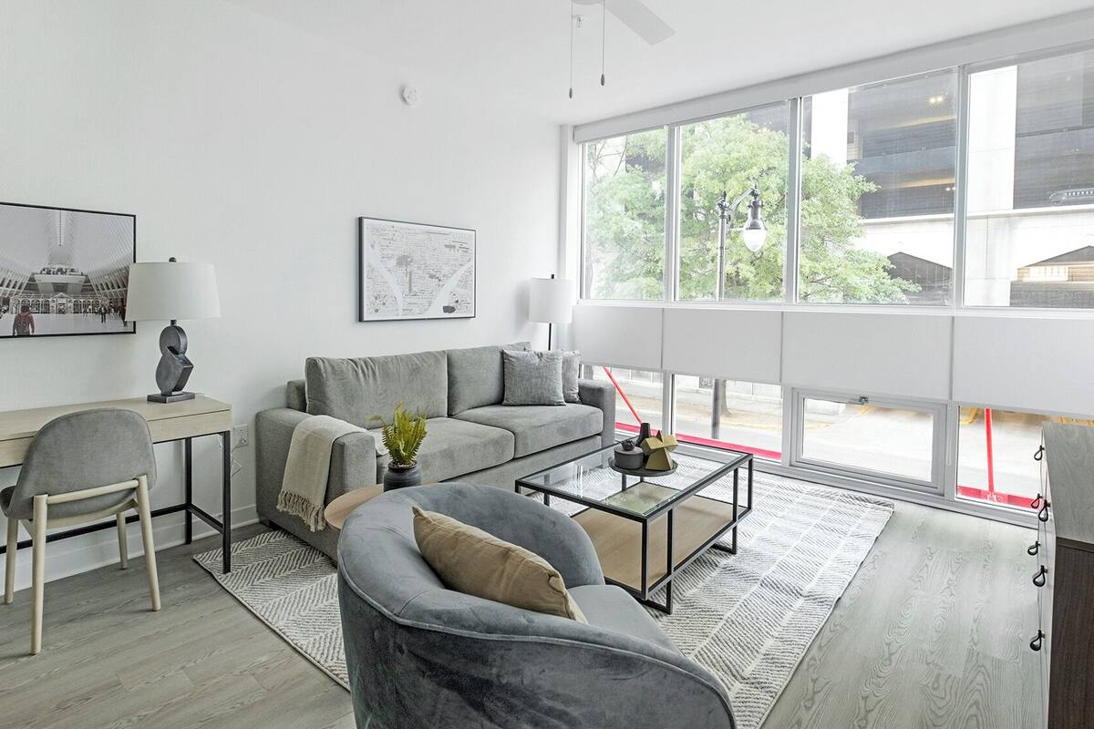 Una sala de estar moderna con sofá, mesa de centro y escritorio. Hay ventanas grandes con vistas a árboles y edificios.