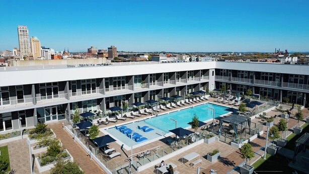 Un área de piscina en la azotea con sillas de descanso y sombrillas, rodeada por un edificio moderno de varios pisos y grandes ventanales.