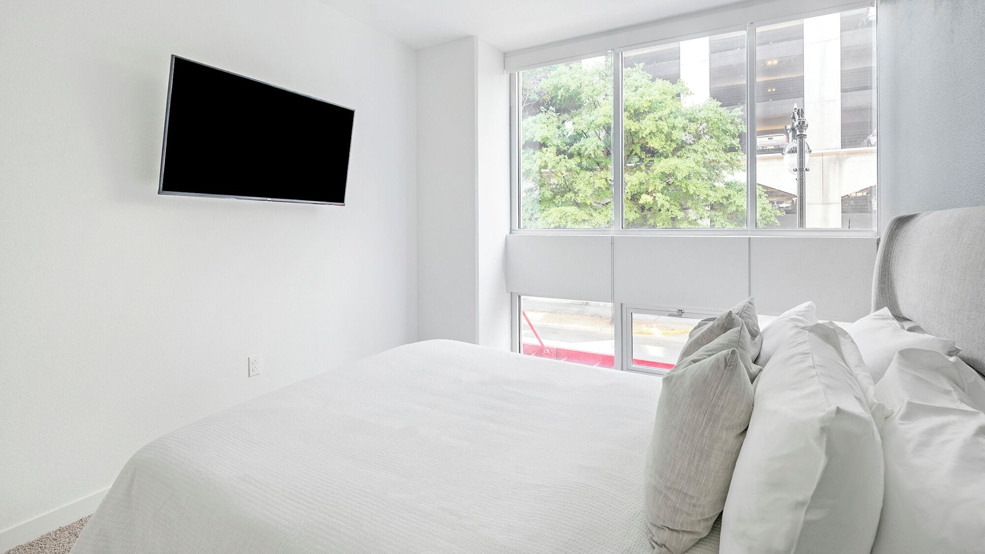 Une chambre à coucher avec un lit, un téléviseur fixé au mur et une fenêtre donnant sur des arbres et des immeubles.