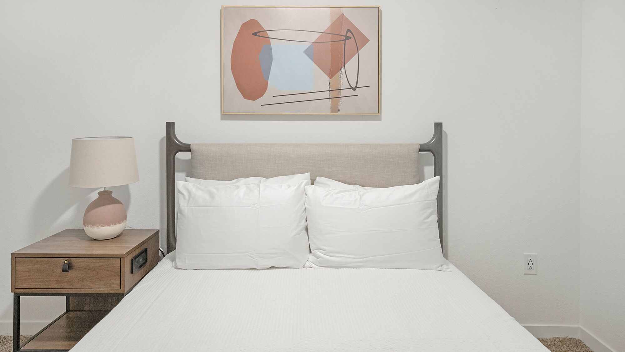 A bed with white bedding, a bedside table with a lamp, and a framed abstract painting on the wall.