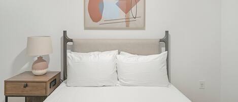 A bed with white bedding, a bedside table with a lamp, and a framed abstract painting on the wall.