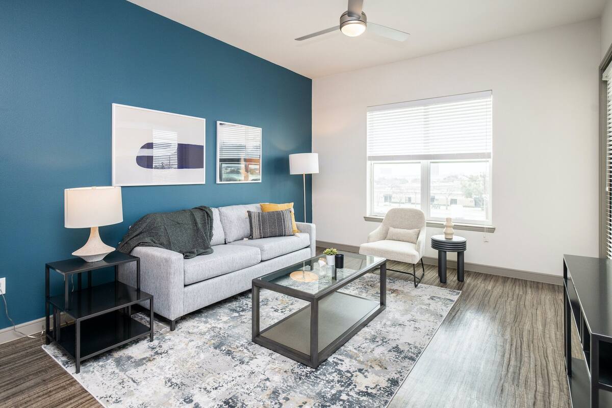 Ein modernes Wohnzimmer mit blauer Akzentwand, einem grauen Sofa, einem weißen Sessel, einem Glastisch und einer Stehlampe.