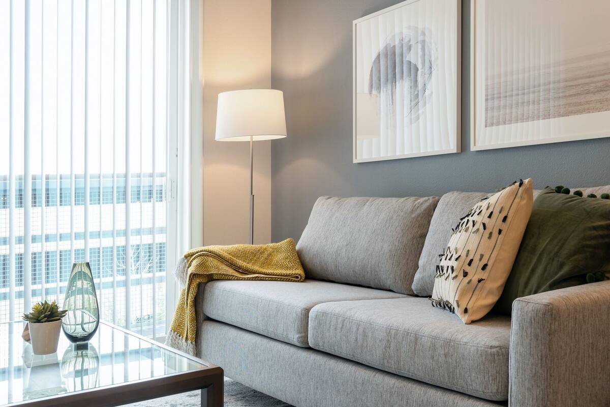 Ein modernes Wohnzimmer mit einem grauen Sofa, einem Glastisch und einer Stehlampe. An der Wand hängen zwei gerahmte Kunstwerke, und ein Fenster mit Jalousien.
