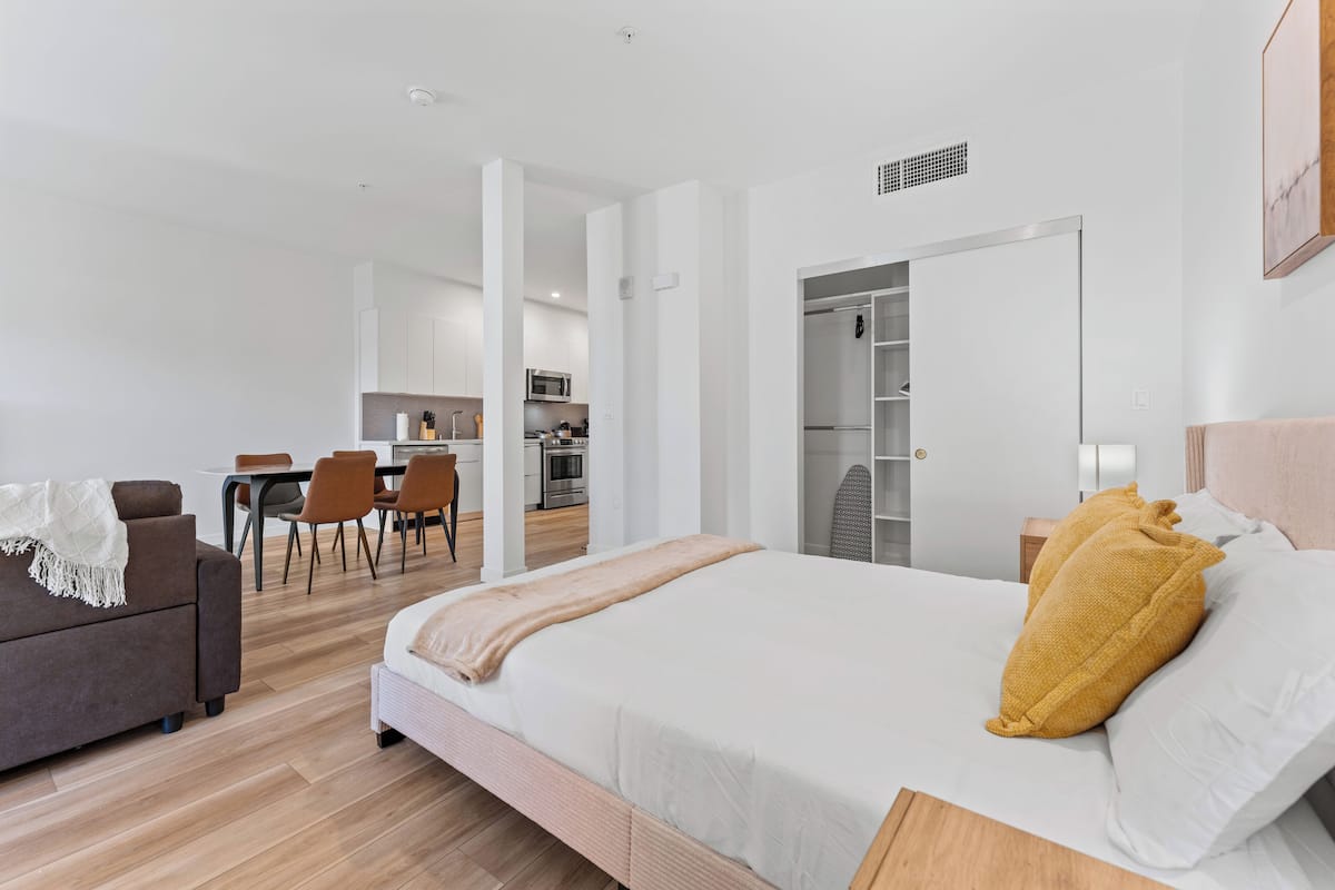 A modern hotel room with a large bed, a sofa, a dining area, and a kitchen in the background.