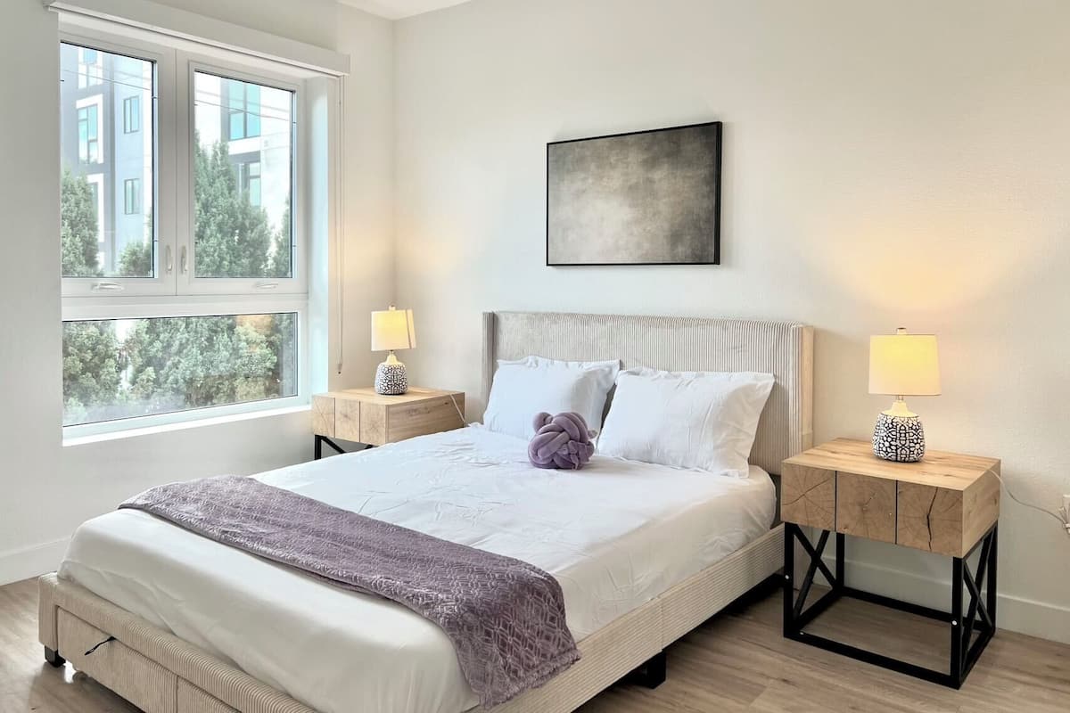 A modern bedroom with a large bed, two bedside tables, and a window with a view of buildings and trees.