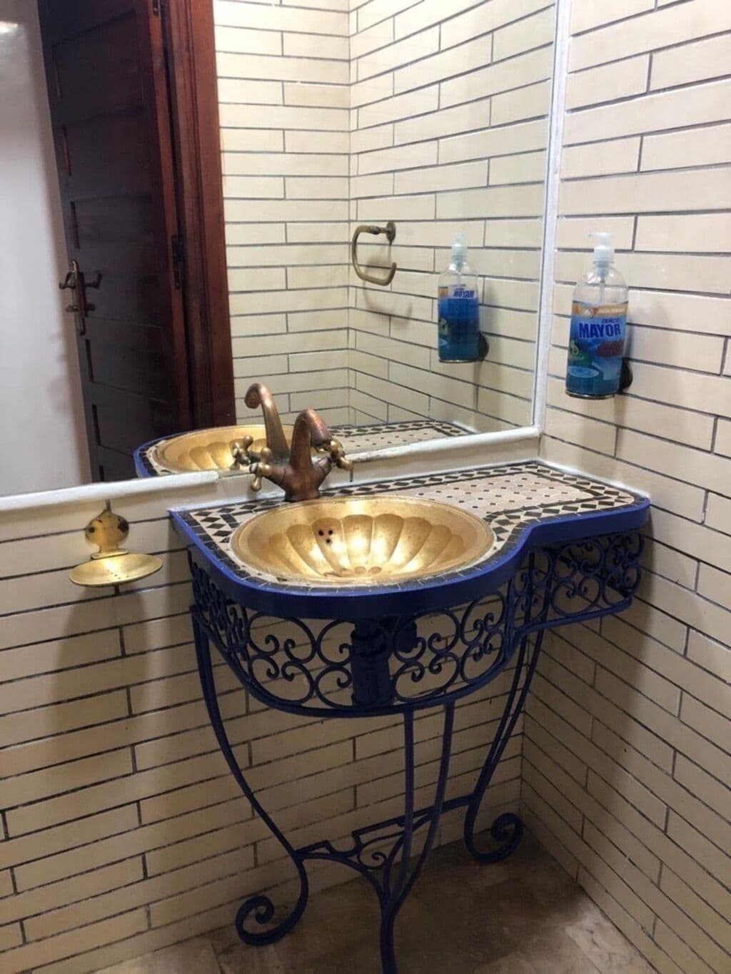 A bathroom with a gold sink, blue decorative metal frame, and a mirror reflecting the room.